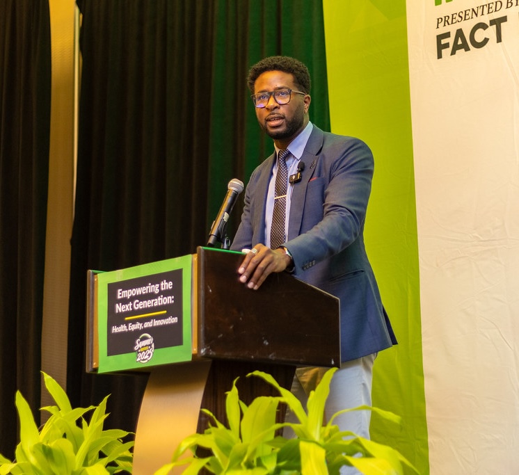 Xavier Ramey speaks at a podium with a microphone. The podium sign reads “Empowering the Next Generation: Health, Equity, and Innovation, Summer Institute 2023.” Green plants are in the foreground, and a banner in the background shows the logo for Fact Forward.